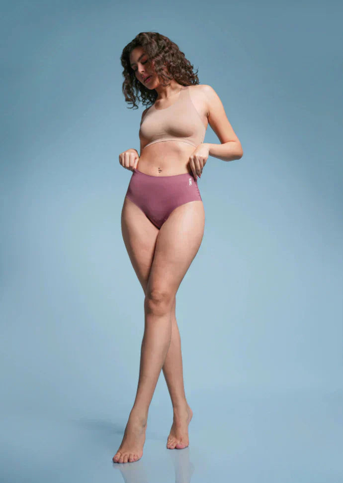 Woman with curly hair wearing a beige  sports bra and pink mid-waist briefs against a light blue background.