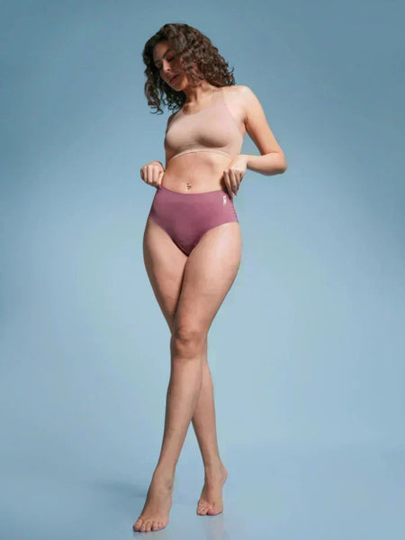 Woman with curly hair wearing a beige  sports bra and pink mid-waist briefs against a light blue background.