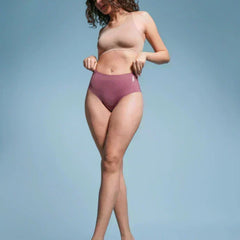 Woman with curly hair wearing a beige  sports bra and pink mid-waist briefs against a light blue background.