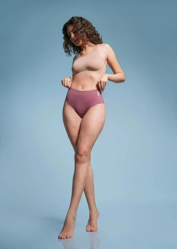 Woman with curly hair wearing a beige  sports bra and pink mid-waist briefs against a light blue background.