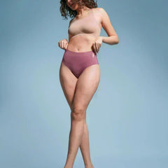 Woman with curly hair wearing a beige  sports bra and pink mid-waist briefs against a light blue background.