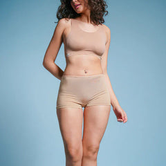 Person with curly hair wearing a beige sports bra and beige boyshort underwear against a blue background.
