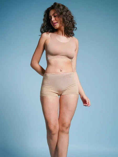 Person with curly hair wearing a beige sports bra and beige boyshort underwear against a blue background.