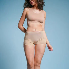 Person with curly hair wearing a beige sports bra and beige boyshort underwear against a blue background.