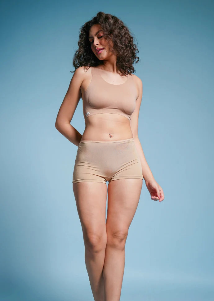 Person with curly hair wearing a beige sports bra and beige boyshort underwear against a blue background.