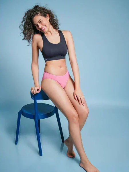 Woman in black sports bra and pink underwear sitting on a blue chair against a light blue background