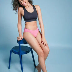 Woman in black sports bra and pink underwear sitting on a blue chair against a light blue background