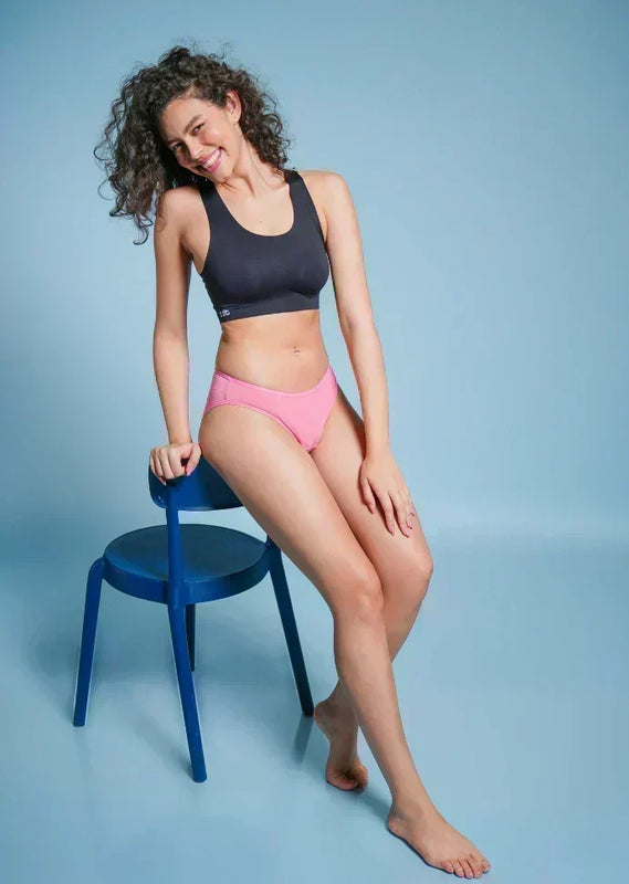 Woman in black sports bra and pink underwear sitting on a blue chair against a light blue background