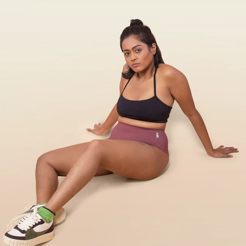 Woman wearing a black sports bra and pink underwear sitting on a beige background