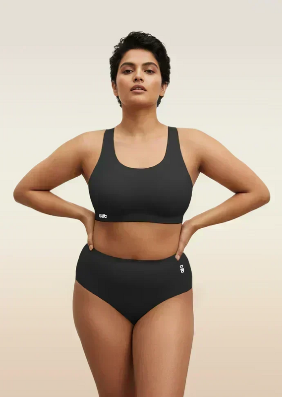 Woman wearing a black sports bra and mid-waist underwear on a beige background