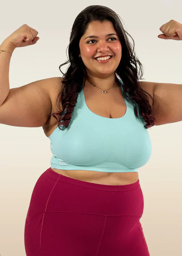 Woman flexing her muscles wearing a light blue bra and maroon leggings on a plain background
