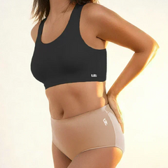 Woman wearing a black sports bra and beige mid-waist briefs, standing at a side angle in warm sunlight.