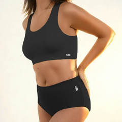 Woman wearing a black sports bra and mid-waist underwear with a visible brand logo on a neutral background
