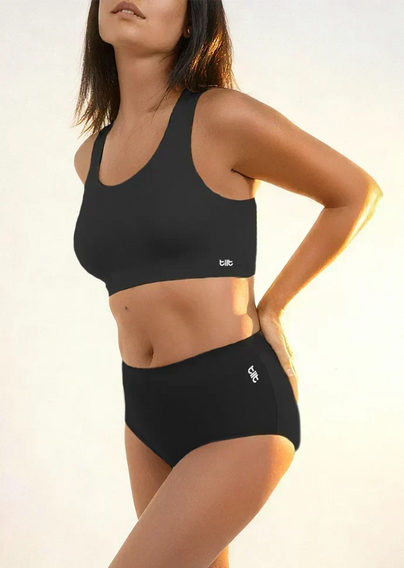 Woman wearing a black sports bra and mid-waist underwear with a visible brand logo on a neutral background