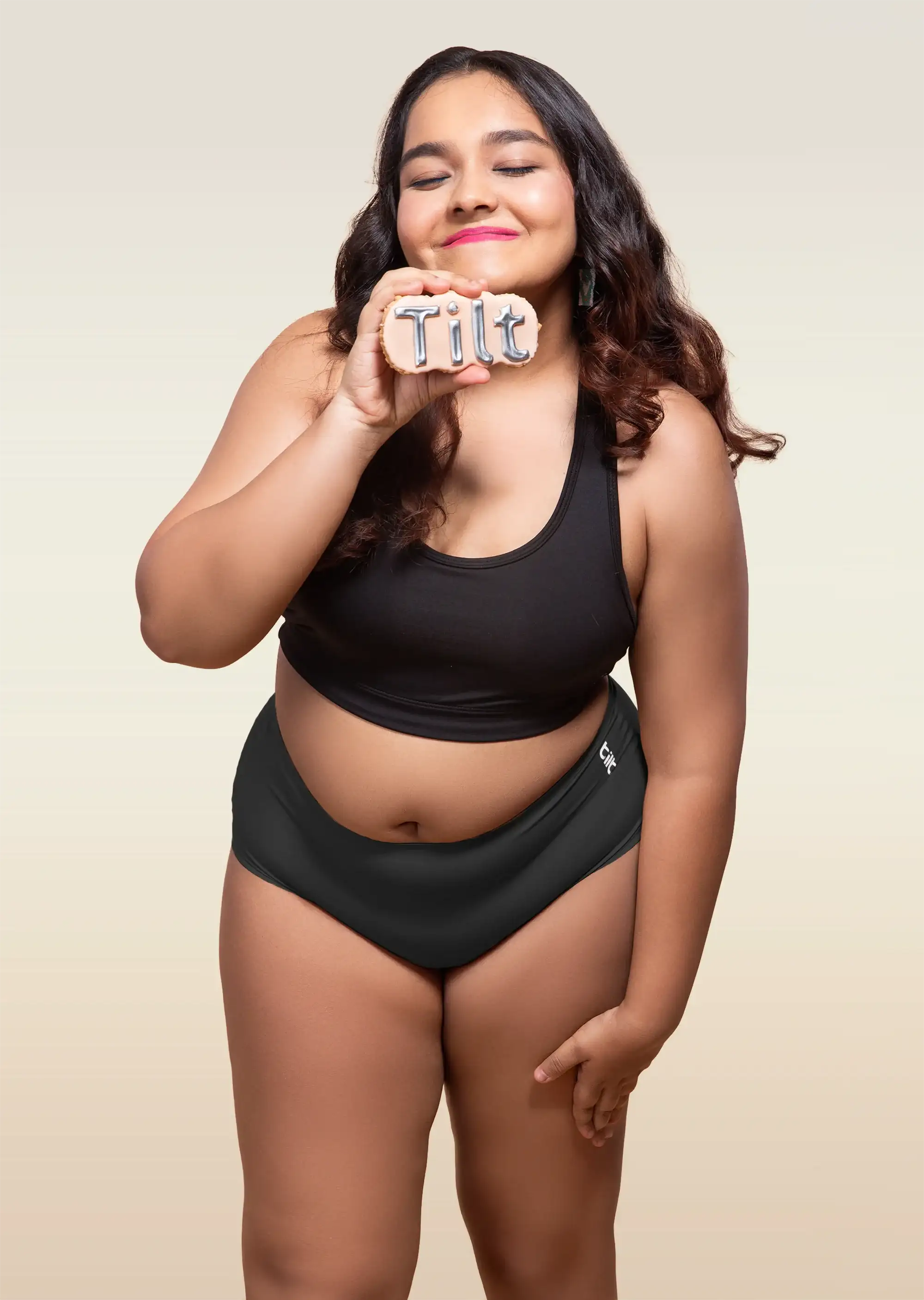 Woman wearing a black underwear holding a 'Tilt' sign against a beige background