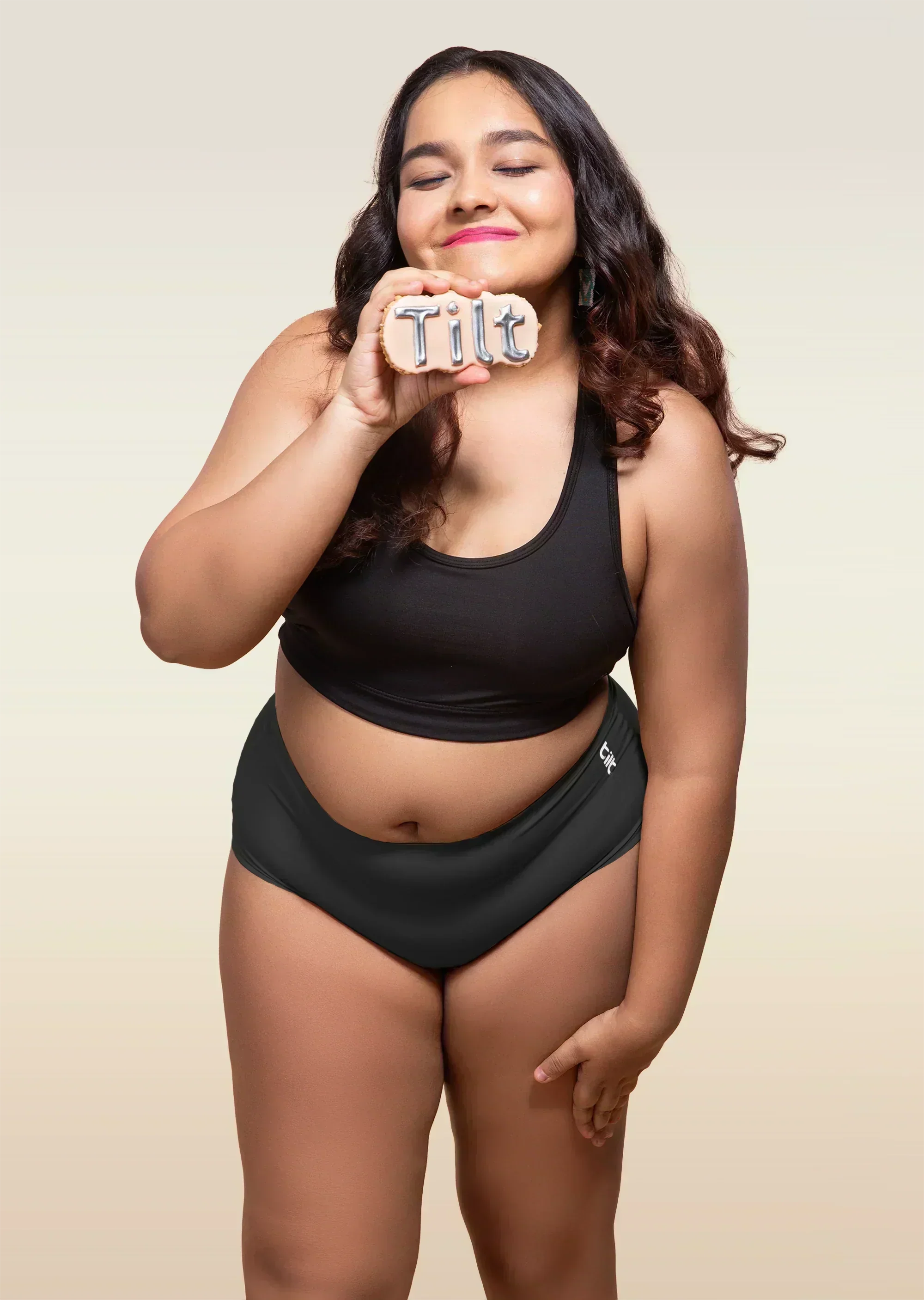 Woman wearing black sports bra and black mid-waist underwear, holding Tilt logo