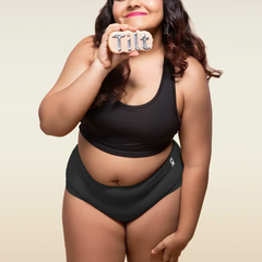Woman wearing black sports bra and black mid-waist underwear, holding Tilt logo