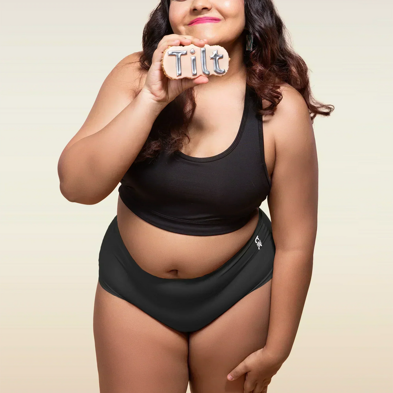 Woman wearing black sports bra and black mid-waist underwear, holding Tilt logo