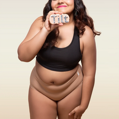 Woman wearing a black tank top and beige mid-waist underwear, holding a 'Tilt' sign.