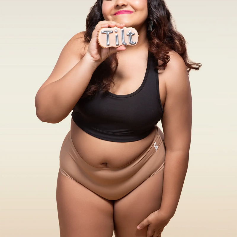 Woman wearing a black tank top and beige mid-waist underwear, holding a 'Tilt' sign.