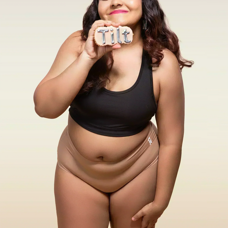 Woman wearing a black tank top and beige mid-waist underwear holding a 'Tilt' sign.