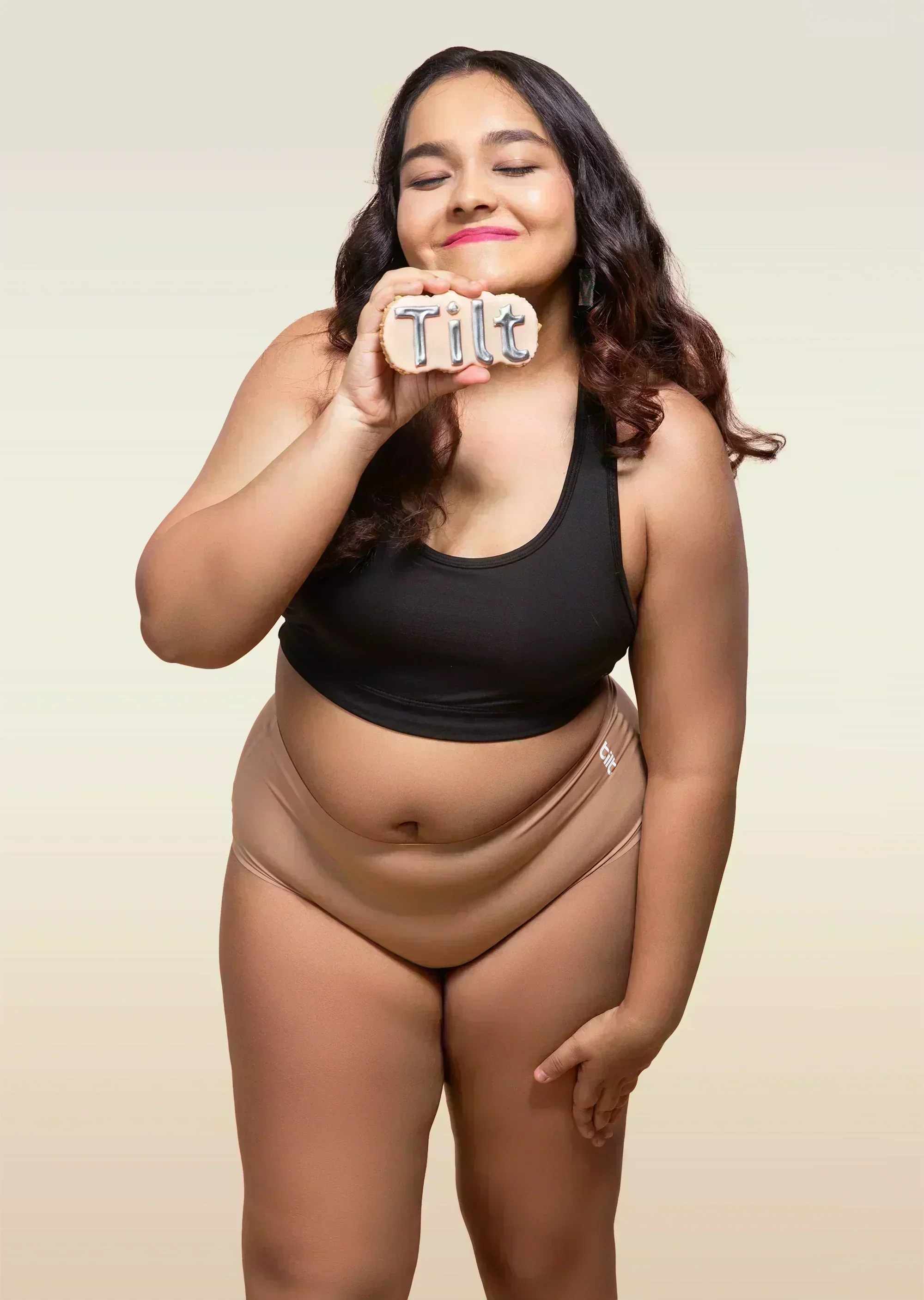 Woman wearing a black tank top and beige mid-waist underwear holding a 'Tilt' sign.