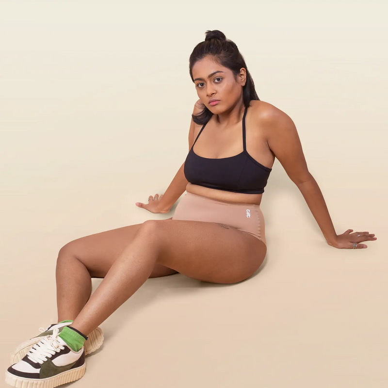 Woman sitting on a beige background wearing a black sports bra and beige underwear.