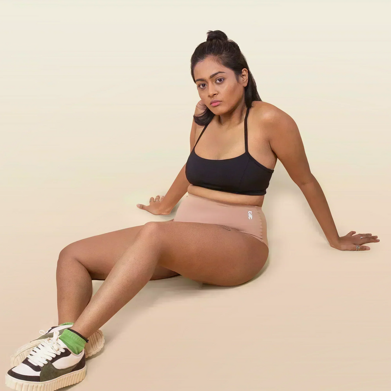 Woman sitting on a beige background wearing a black sports bra and beige underwear.