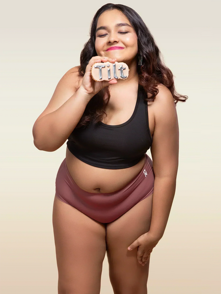Woman wearing a black tank top and pink mid-waist underwear holding a 'Tilt' sign.