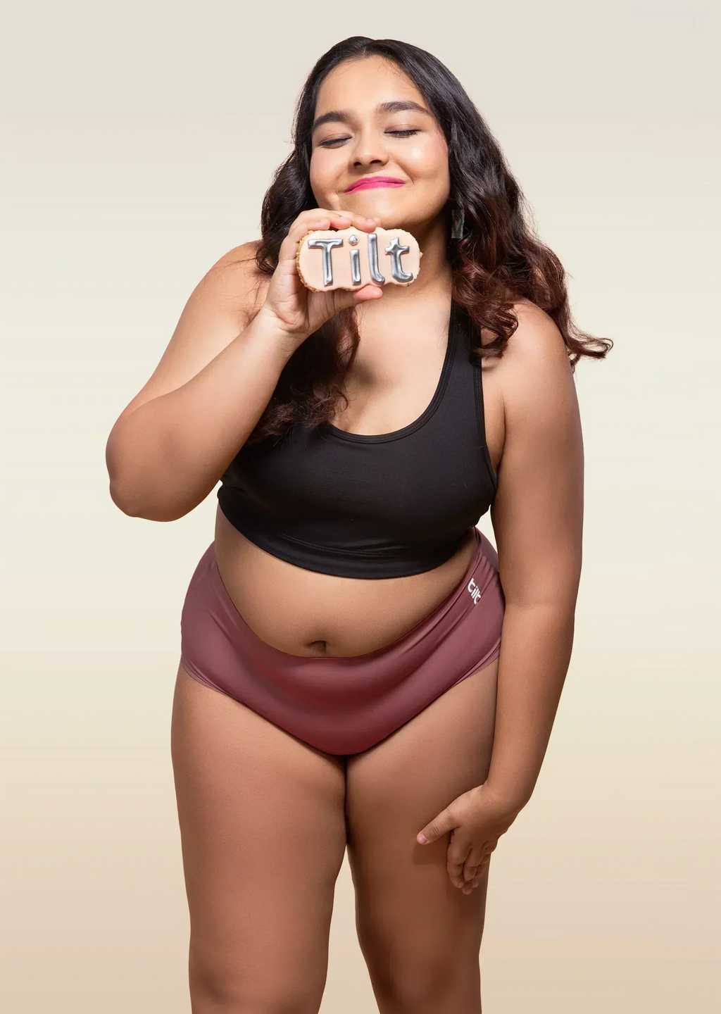 Woman wearing a black tank top and pink mid-waist underwear holding a 'Tilt' sign.