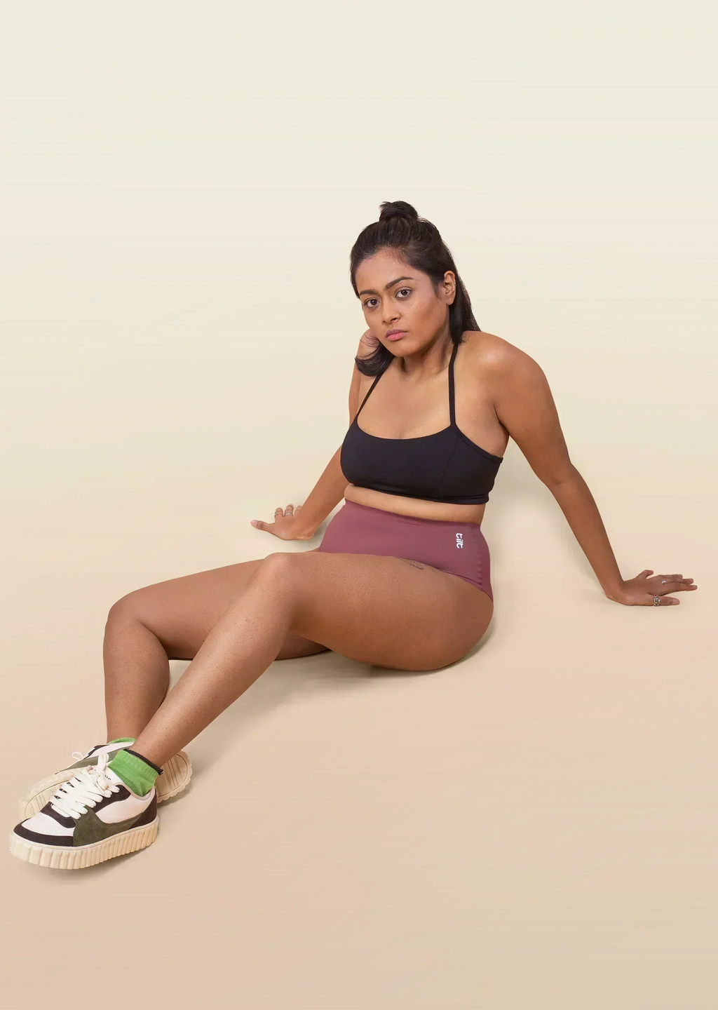 Woman wearing a black sports bra and pink underwear sitting on a beige background