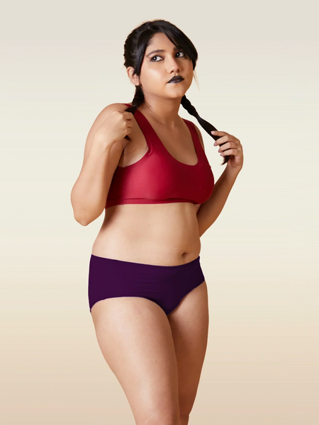 Woman wearing a red sports bra and violet mid-waist underwear against a beige background
