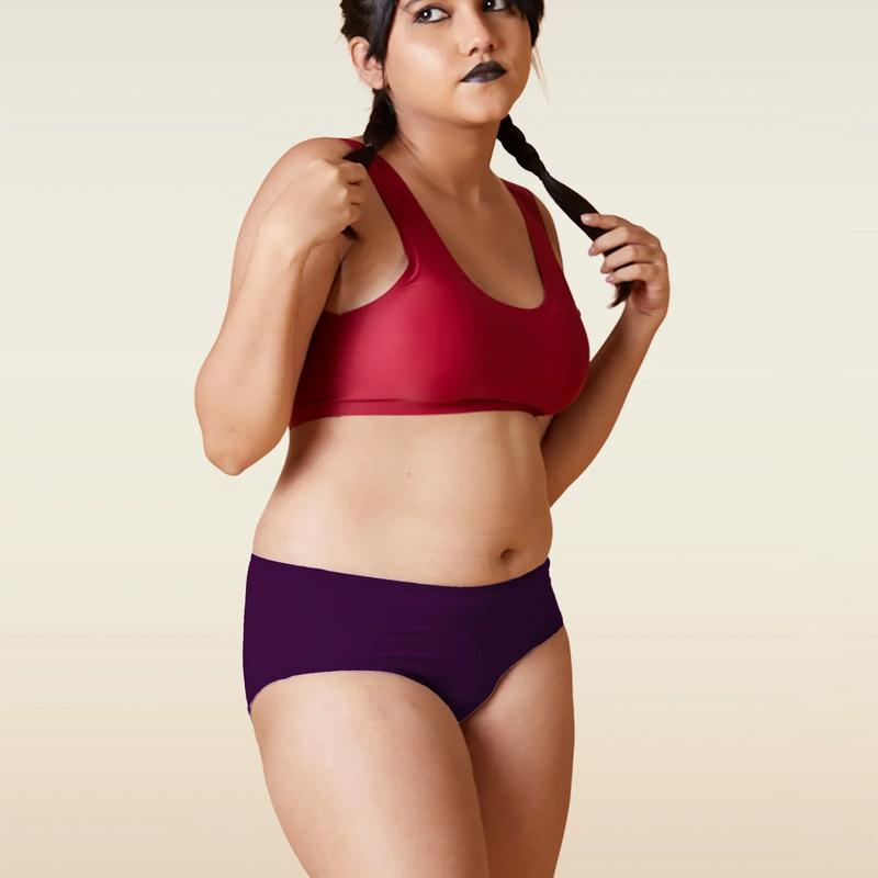 Woman wearing a red sports bra and violet mid-waist underwear against a beige background