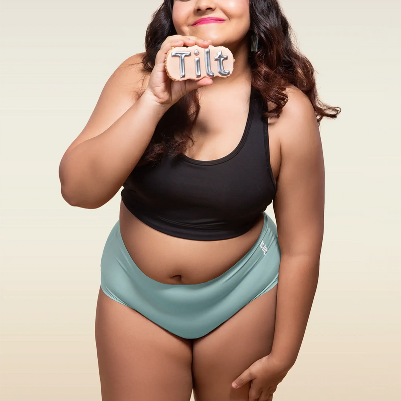 Woman wearing a black top and blue mid-waist underwear holding a sign that says 'Tilt'.