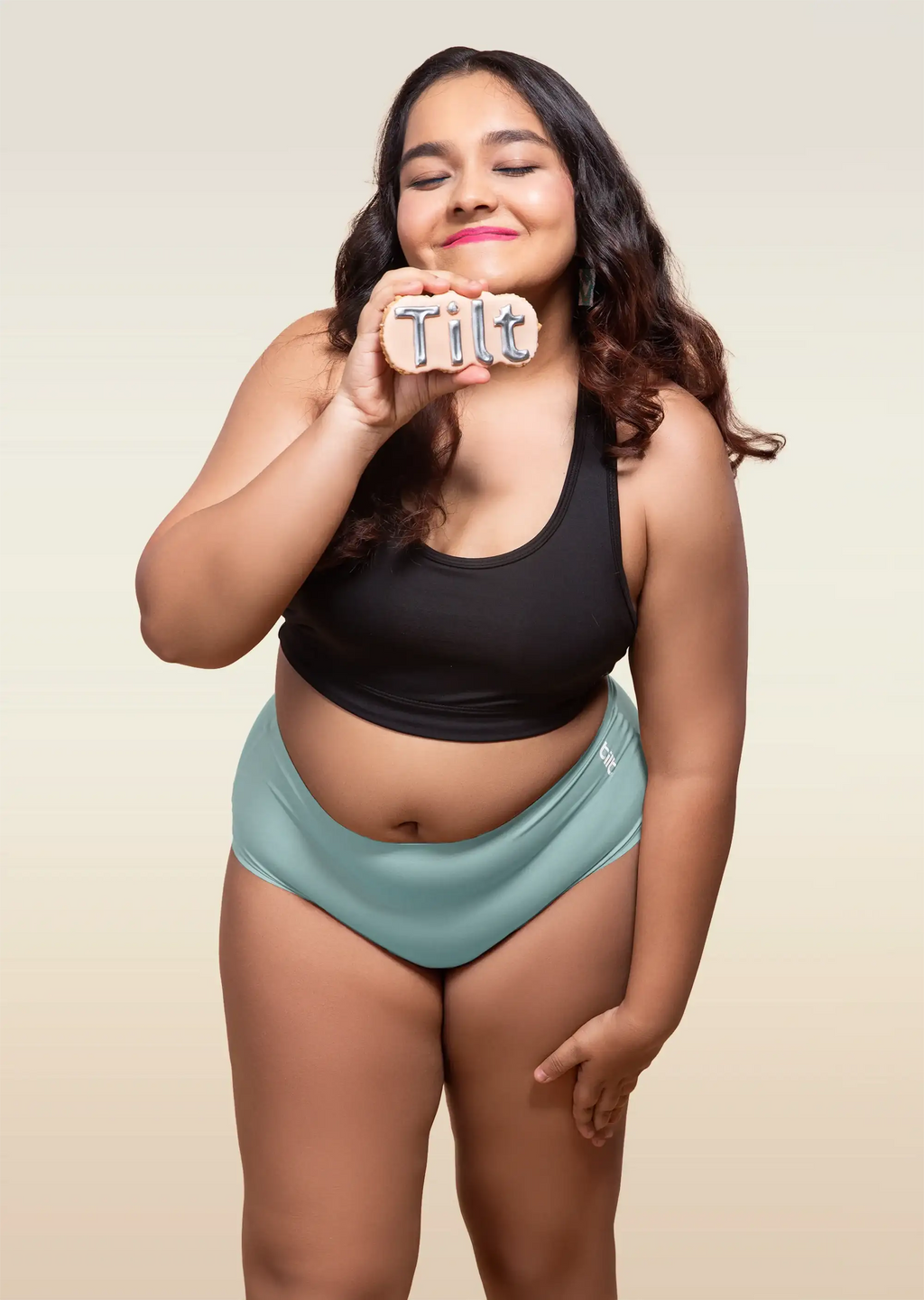 Woman wearing a black top and blue mid-waist underwear holding a sign that says 'Tilt'.
