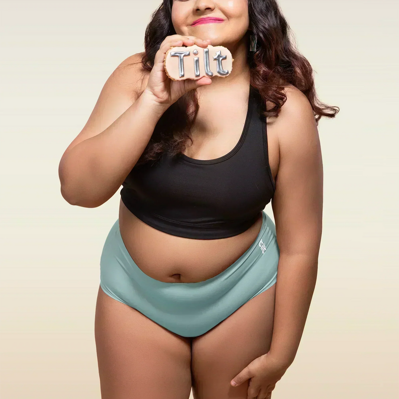 Woman wearing black sports bra and blue mid-waist seamless underwear holding a 'Tilt' sign.
