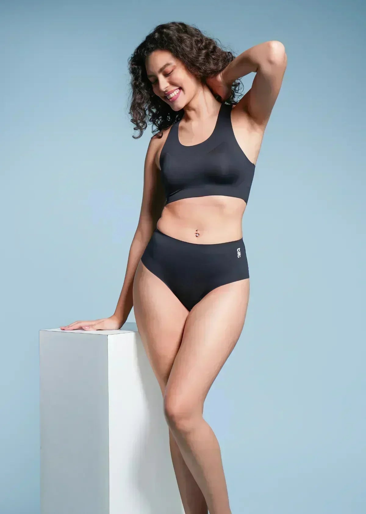 Woman wearing a black sports bra and mid-waist underwear against a light blue background