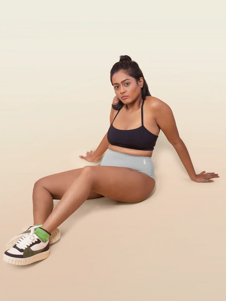 Woman sitting on a beige background wearing a black sports bra and grey underwear.