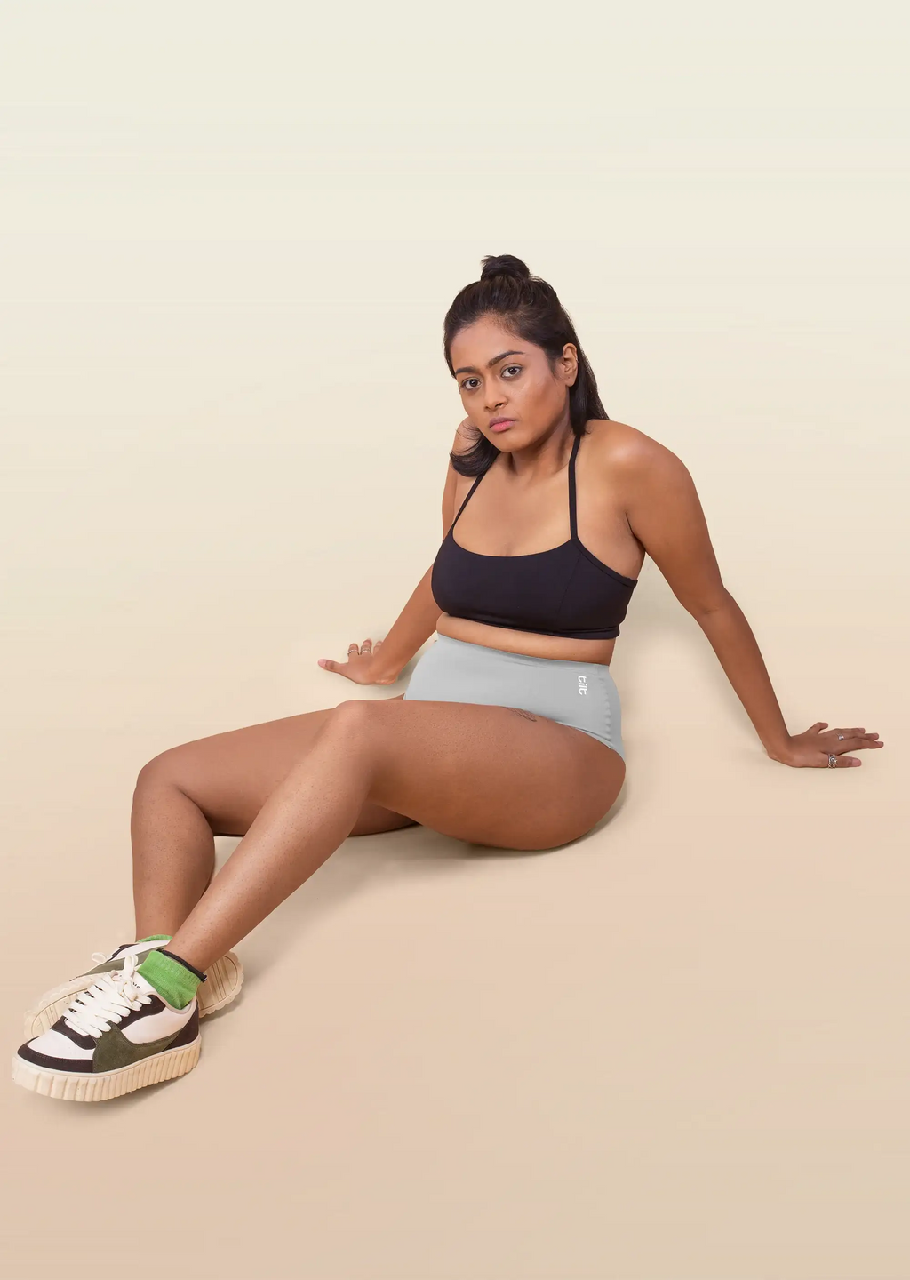 Woman sitting on a beige background wearing a black sports bra and grey underwear.