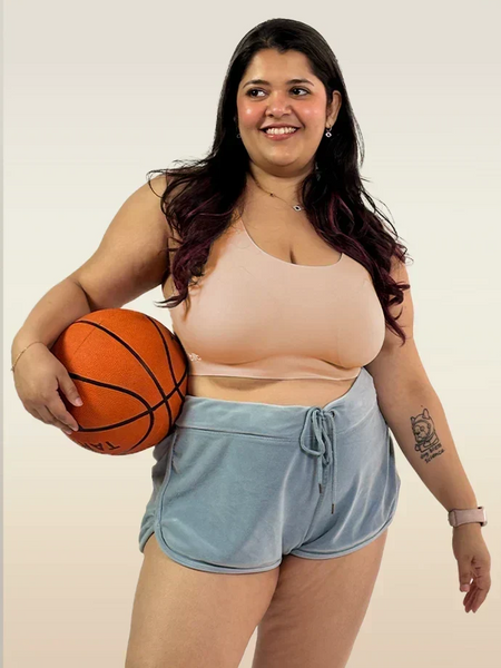 Person with long dark hair smiling and holding a basketball while wearing a beige sports bra and light blue velvet-style shorts.