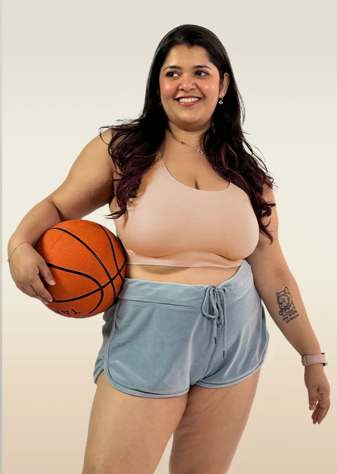 Person with long dark hair smiling and holding a basketball while wearing a beige sports bra and light blue velvet-style shorts.