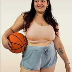 Person with long dark hair smiling and holding a basketball while wearing a beige sports bra and light blue velvet-style shorts.